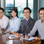Group of four business people sitting at desk in modern office, holding meeting, smiling at camera. Business team concept